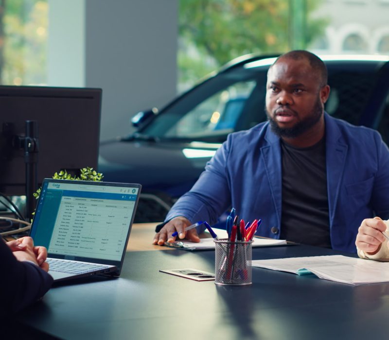 Dealership salesman looking over appointment list on laptop spreadsheet to schedule car test drive for customers. Showroom agent discussing with clients, organizing test drive, camera A